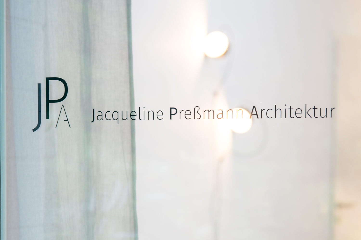 Blick von außen durch die Glastür in das Architekturbüro. Der Schriftzug „JPA Jacqueline Preßmann Architektur“ steht im Fokus, während unscharfe Wandleuchten im Hintergrund eine ruhige Atmosphäre schaffen.