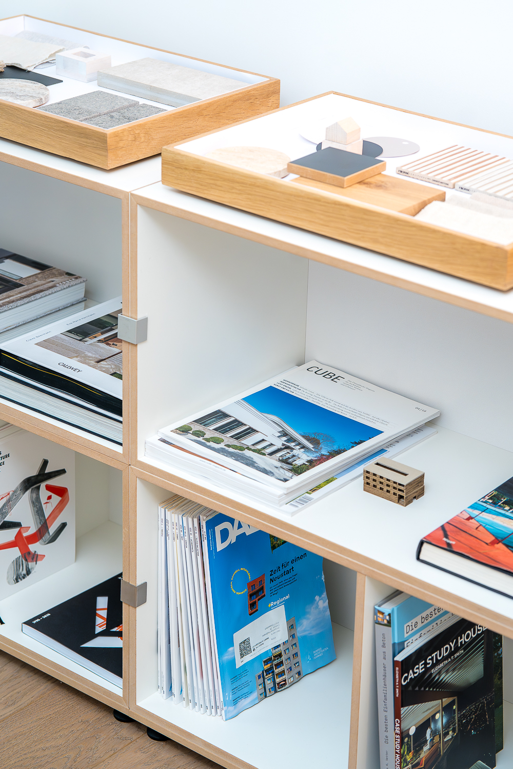Regal im Architekturbüro mit Materialbibliothek, Architekturzeitschriften und Fachbüchern. Materialtabletts mit Holz-, Stein- und Farbmustern vermitteln einen Einblick in die tägliche Entwurfsarbeit.