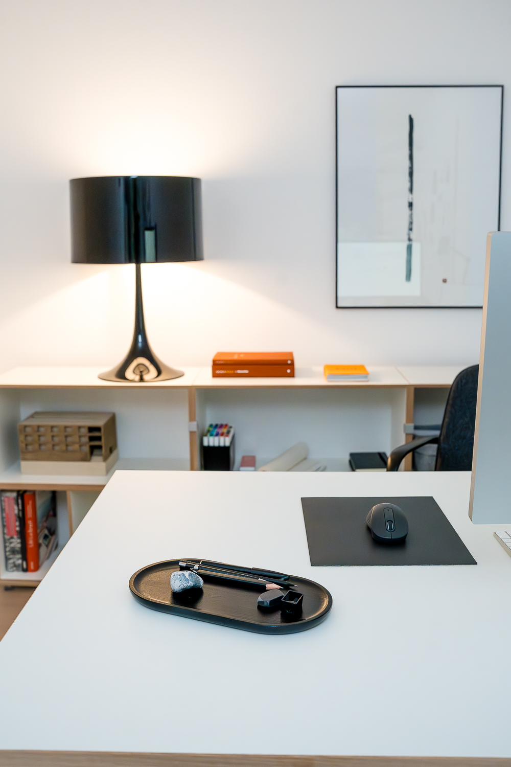 Detailaufnahme des Arbeitsplatzes im Architekturbüro. Minimalistisch gestalteter Schreibtisch mit sorgfältig ausgewählten Arbeitsutensilien, im Hintergrund Architekturmodell, Fachliteratur und Tuschezeichnung in einer ruhigen, reduzierten Arbeitsumgebung.