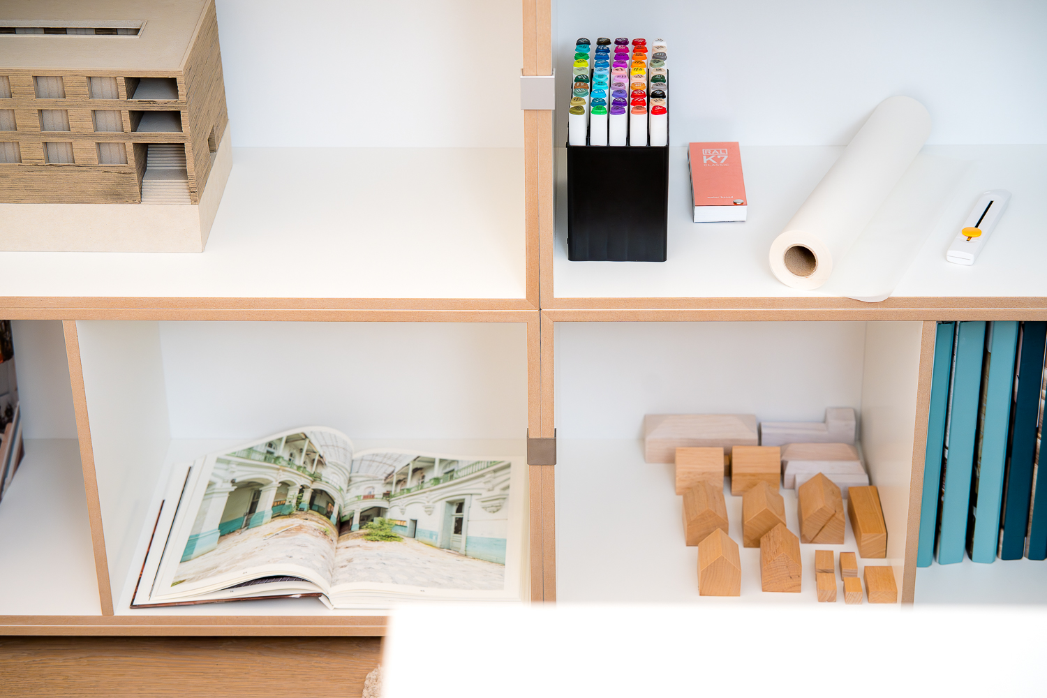 Detailaufnahme eines Regals im Architekturbüro mit Architekturmodell, Zeichenwerkzeugen, Skizzenpapier, Holzmodellen und Fachliteratur. Die sorgfältig zusammengestellten Arbeitsmaterialien veranschaulichen die tägliche Entwurfsarbeit.