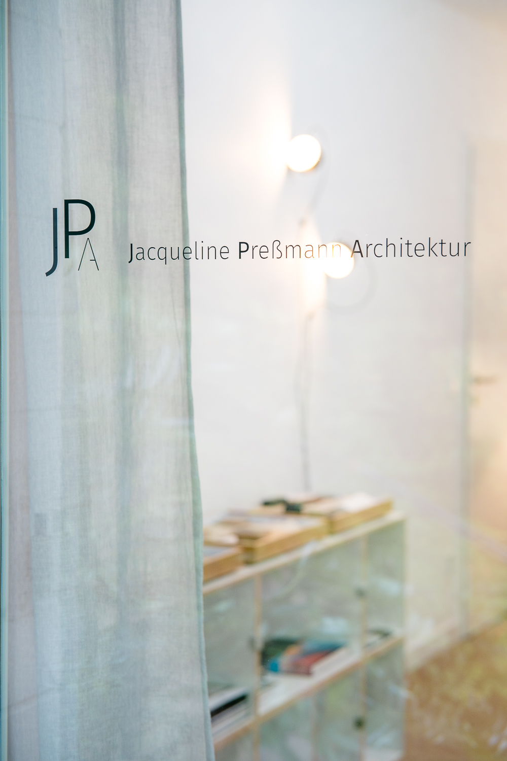Blick von außen durch die Glastür in das Architekturbüro. Der Schriftzug „JPA Jacqueline Preßmann Architektur“ steht im Fokus, dahinter zeichnen sich Sideboard, Materialien, Bücher und Wandleuchten in einer ruhigen Unschärfe ab