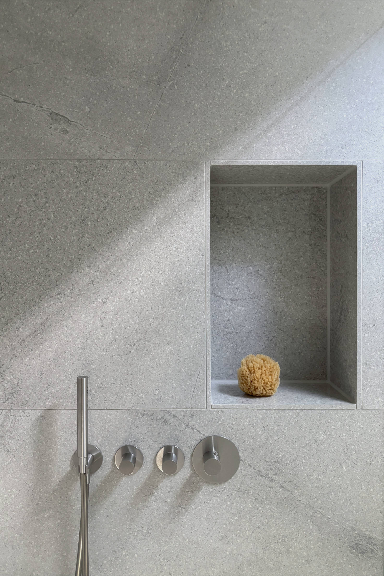 Bad 2: Detail der Dusche mit großzügiger Wandnische aus großformatigem Feinsteinzeug, Edelstahlarmaturen und einem Naturschwamm in der Nische, Tageslicht durch das Oberlicht betont die Materialität der Oberfläche