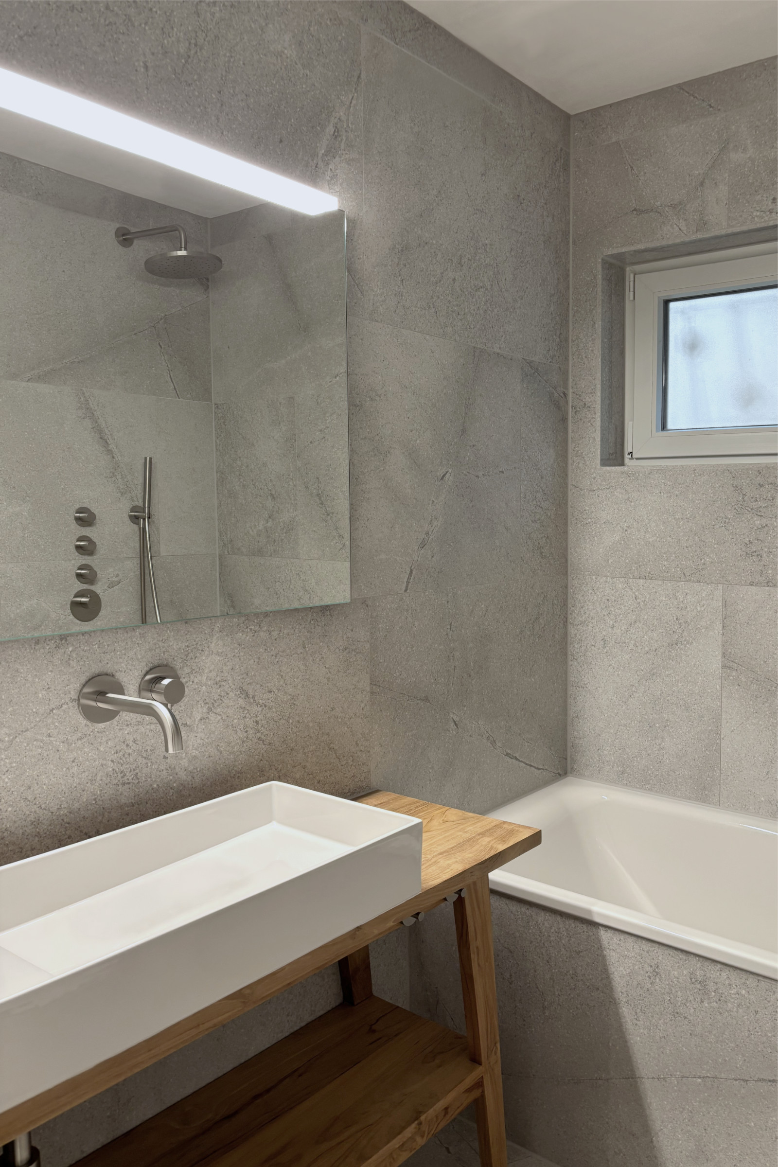Bd 2: Badezimmer mit Emaille Badewanne, großformatigem Feinsteinzeug an Boden und Wänden, Holzwaschtisch mit rechteckigem Aufsatzwaschbecken und Edelstahlarmaturen, natürliches Licht durch Fenster