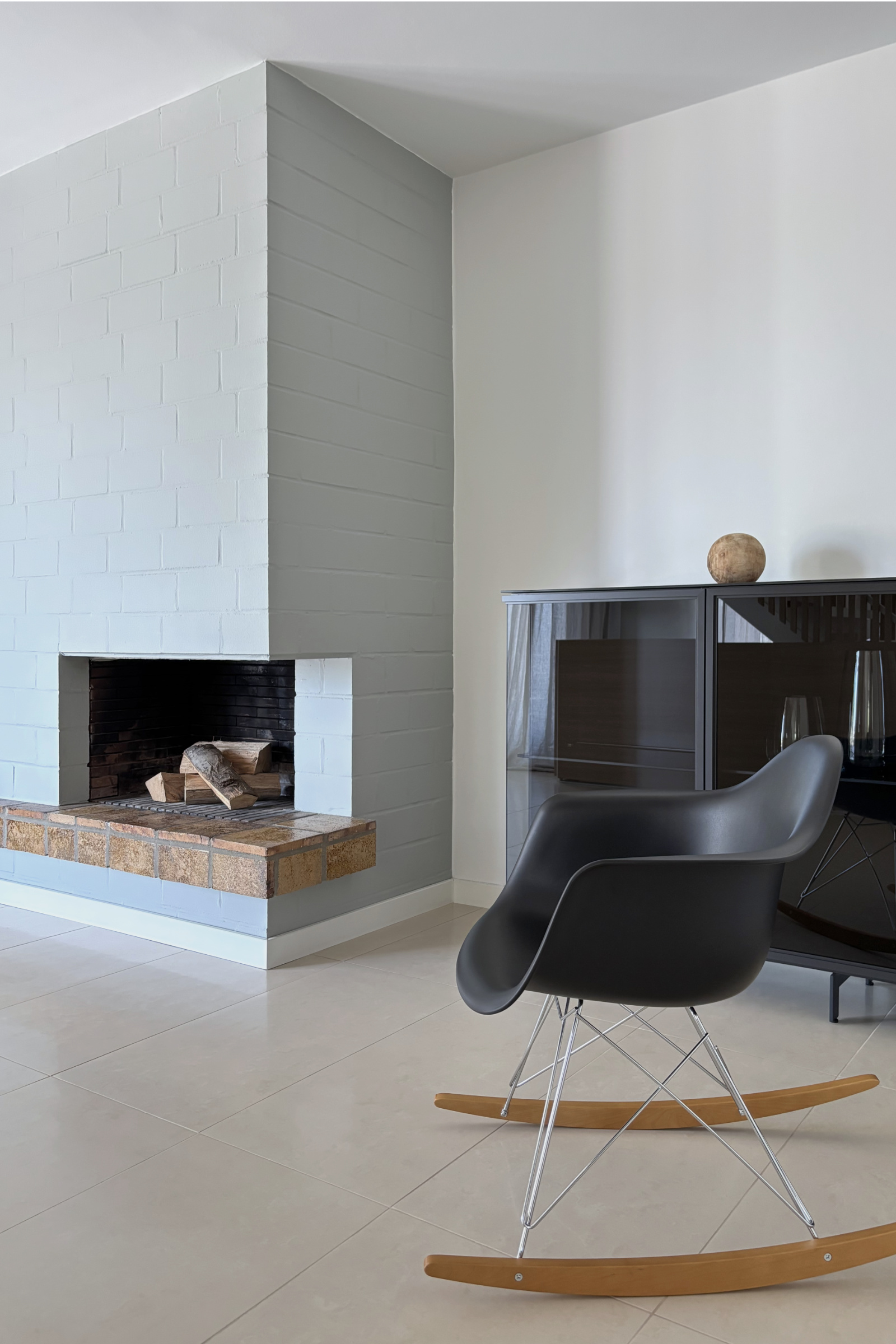 Detailaufnahme mit Kamin aus Sichtmauerwerk, reduziertem Sideboard aus schwarzem Aluminium und Glas sowie schwarzem Rocking chair mit Holzkufen. Die Kombination aus Holz, Metall und mineralischen Oberflächen prägt die Materialwirkung des Raumes