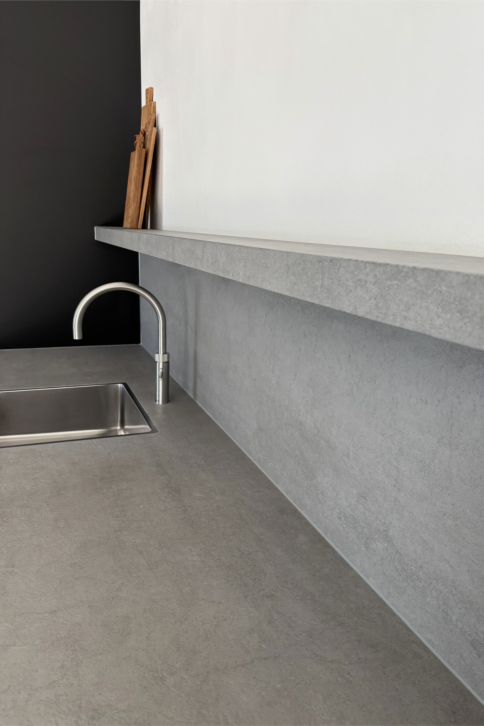 Detailaufnahme der Küchenzeile mit Edelstahlspüle und Armatur. Großformatiges graues Feinsteinzeug bildet Arbeitsplatte, Rückwand und Ablageboard aus einem durchgängigen Materialkonzept