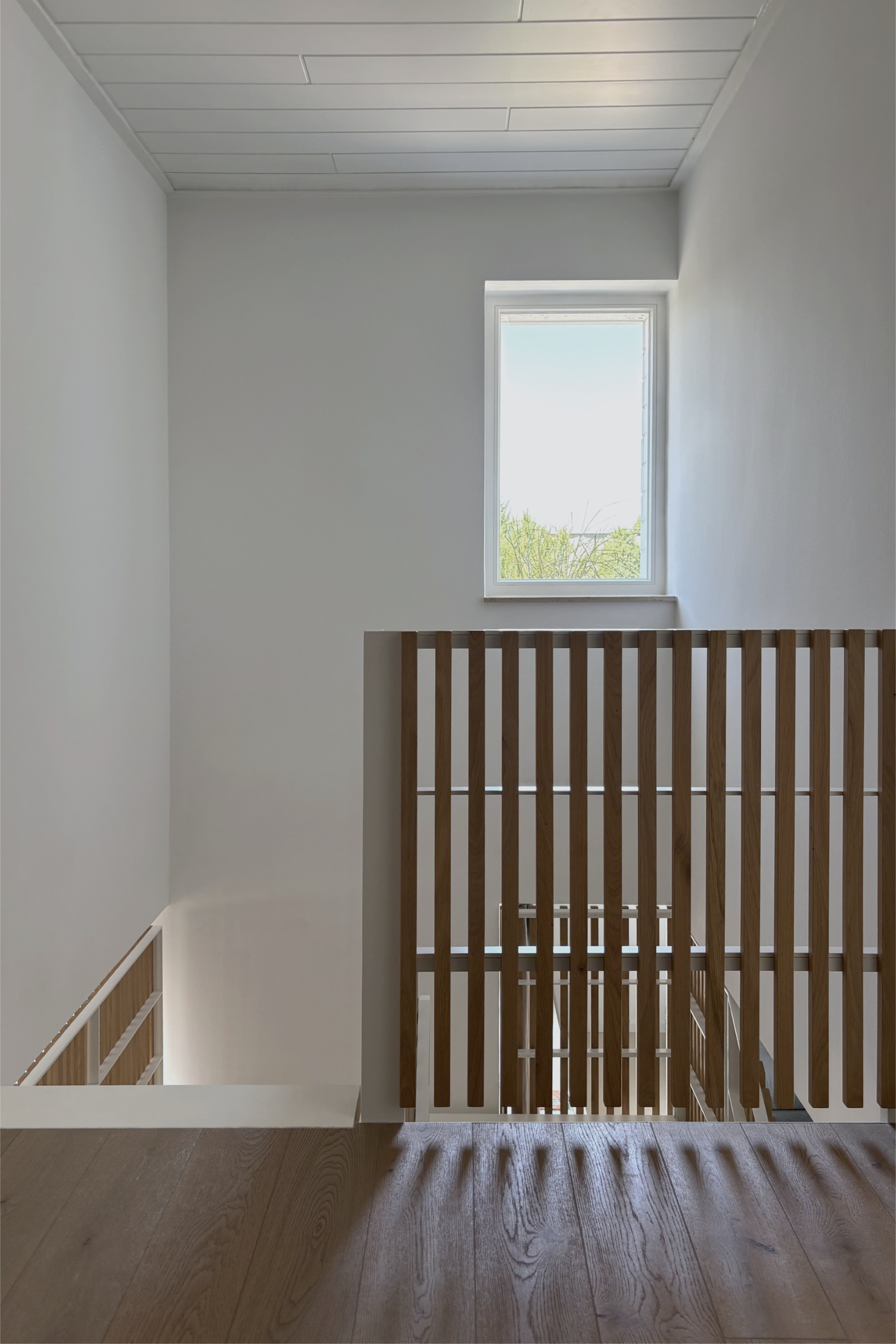Treppe: Blick vom Obergeschoss auf die Brüstung der Treppe mit weißem Stahlgeländer und vertikalen Eichenholzlamellen. Natürliches Tageslicht durch das Fenster erzeugt ein Schattenspiel auf dem Holzboden