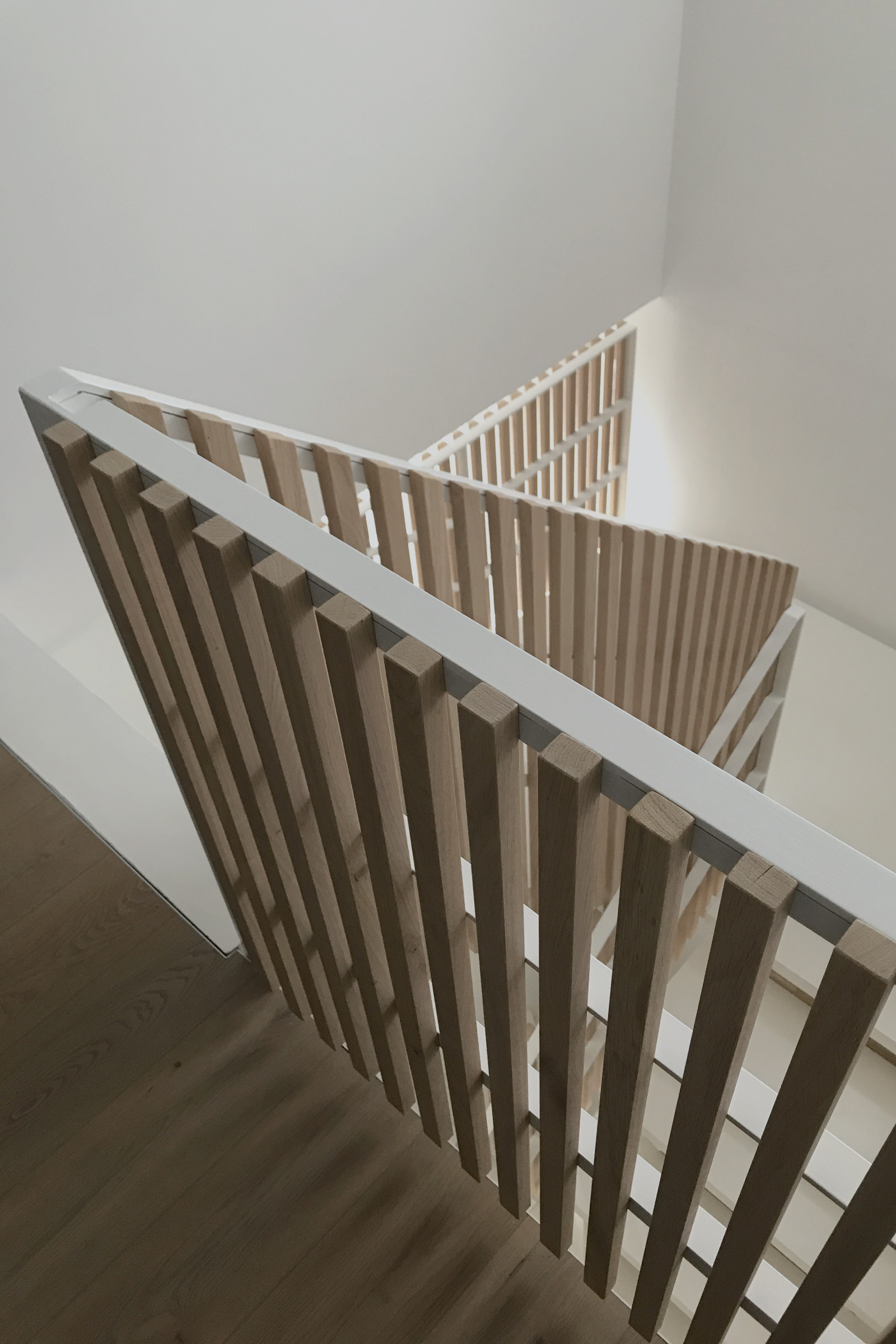 Treppe: Blick schräg über die Treppenbrüstung mit weißem Stahlgeländer und vertikalen Lamellen aus Eichenholz. Die offene Konstruktion macht die räumliche Tiefe des Treppenhauses sichtbar, während Tageslicht Schatten auf den Holzboden zeichnet