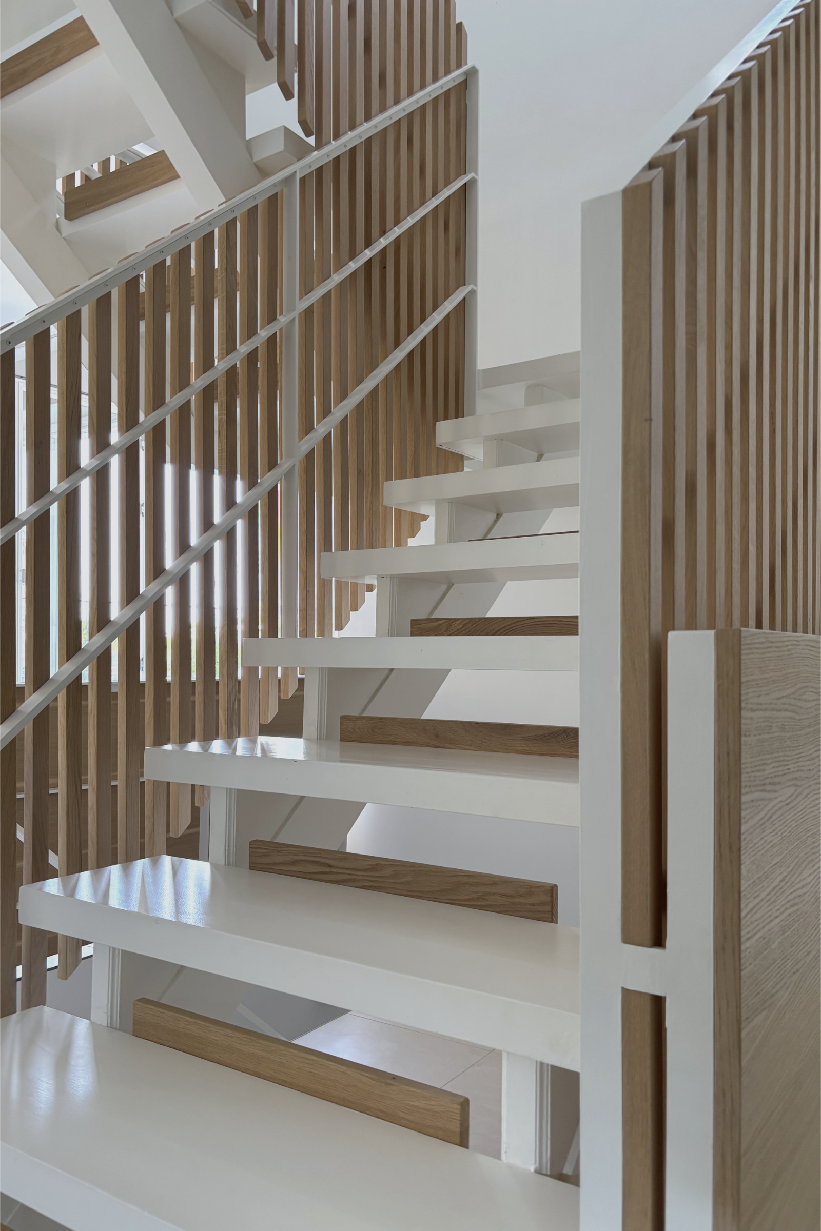 Treppe: Blick auf den offenen Treppenlauf mit weißem Stahlgeländer und vertikalen Eichenholzlamellen. Weiße Holzstufen mit Eichenholz-Setzstufen und natürliches Tageslicht betonen die klare Material- und Formensprache