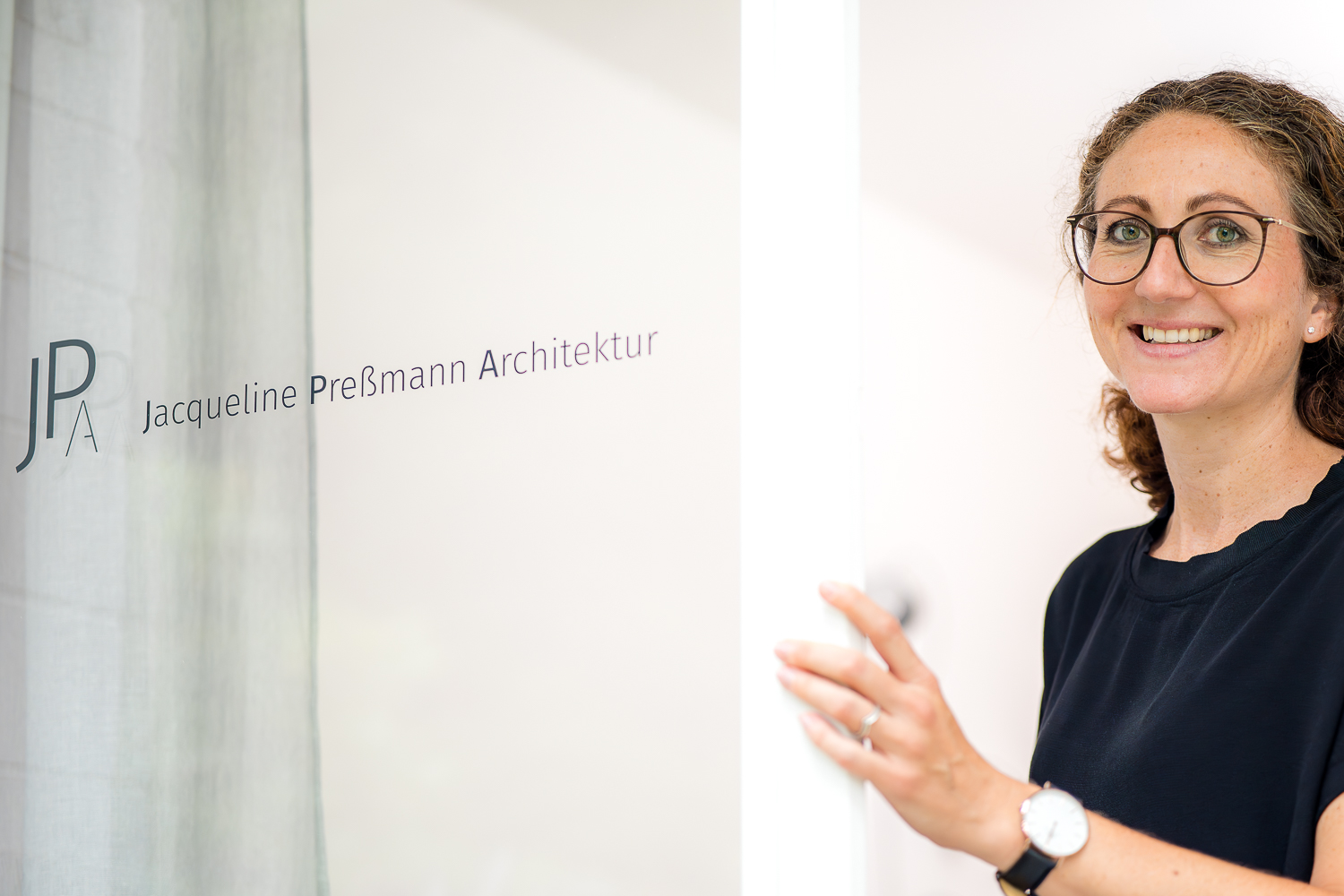 Architektin Jacqueline Preßmann begrüßt Besucherinnen und Besucher im Eingangsbereich ihres Architekturbüros, zu sehen ist der reduzierte Firmenschriftzug JPA Jacqueline Preßmann Architektur auf der Glaseingangstür